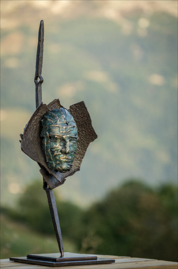 Vu en pied 3/4 droit de la sculpture en bronze et acier Masque, paysage flou dans le fond
